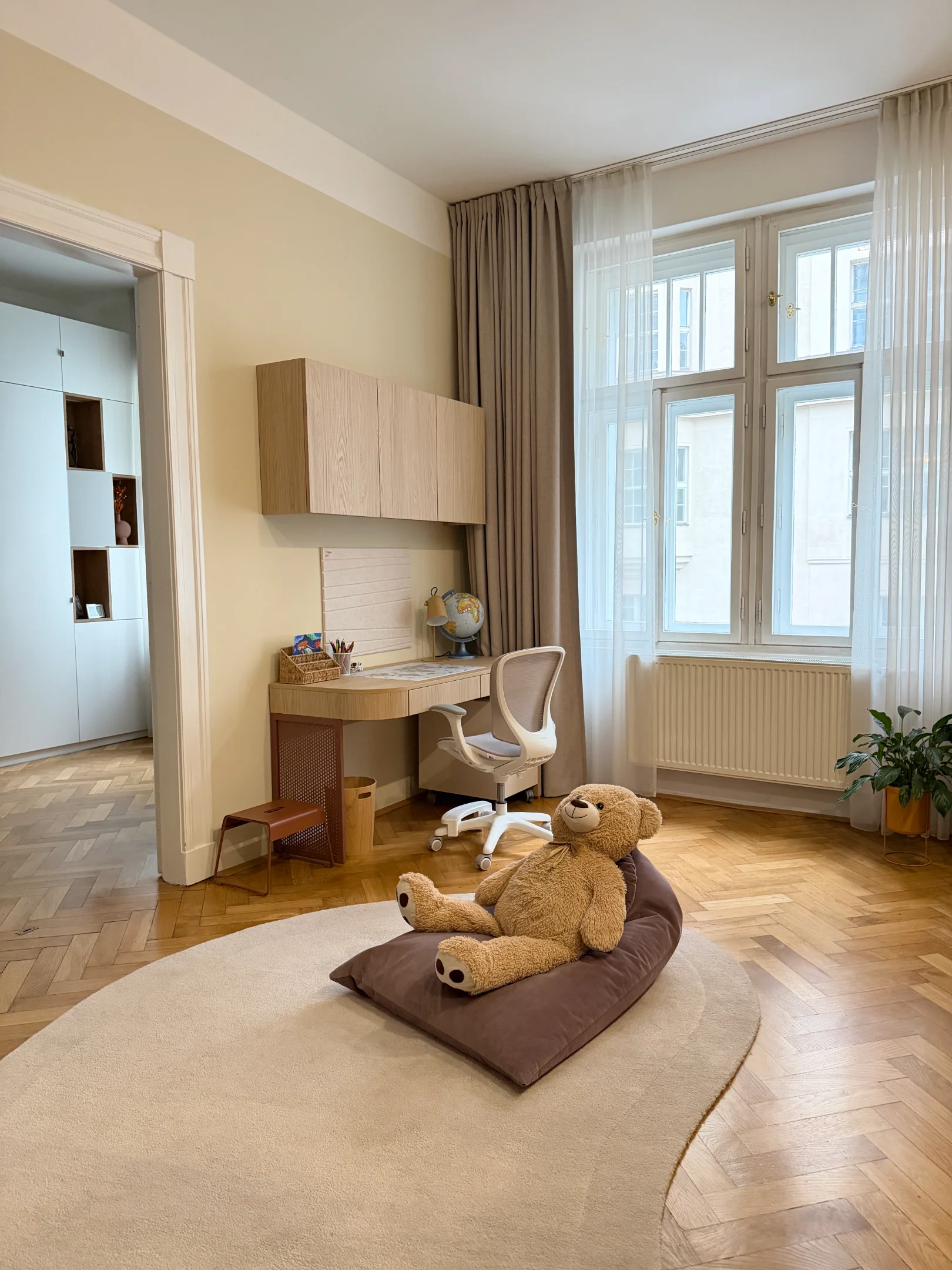 Dětský pokoj s psacím stolem, oknem, kobercem a medvídkem.